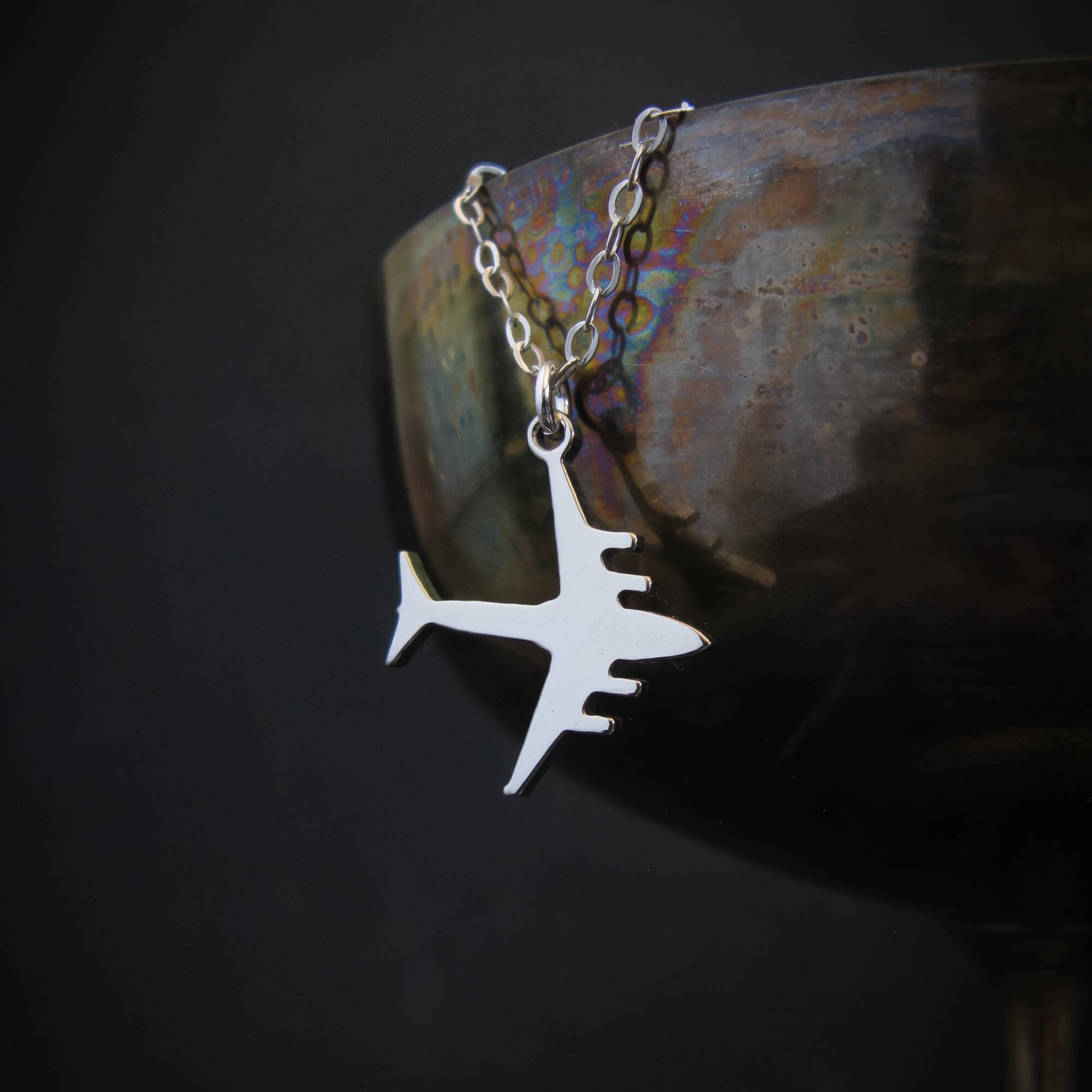 C-17 Globemaster military airplane pendant necklace in sterling silver, draped over the rim of an iridescent metal wine glass against a dark background.