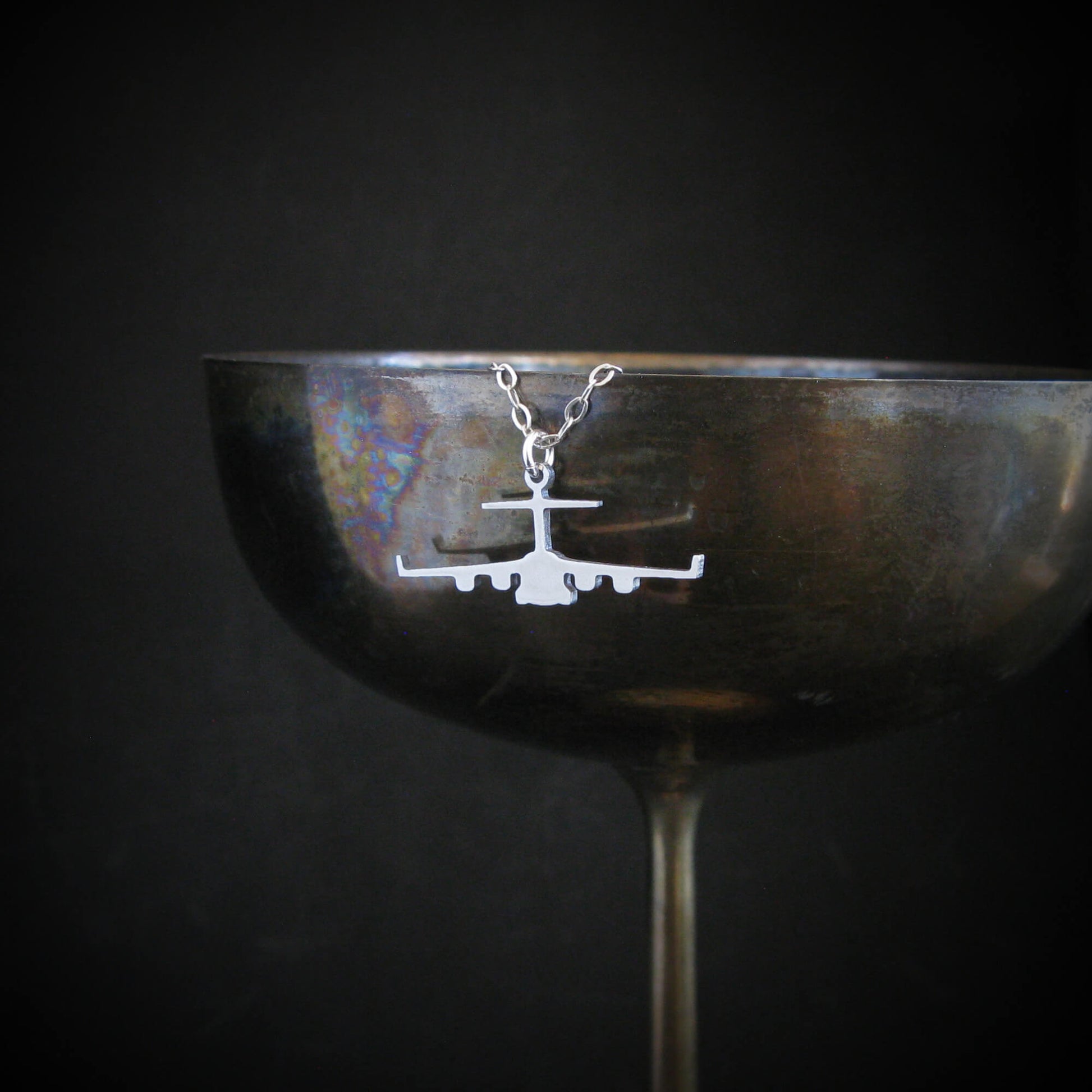 Aviation-themed C-17 aircraft necklace in silver, draped stylishly over a vintage wine glass rim