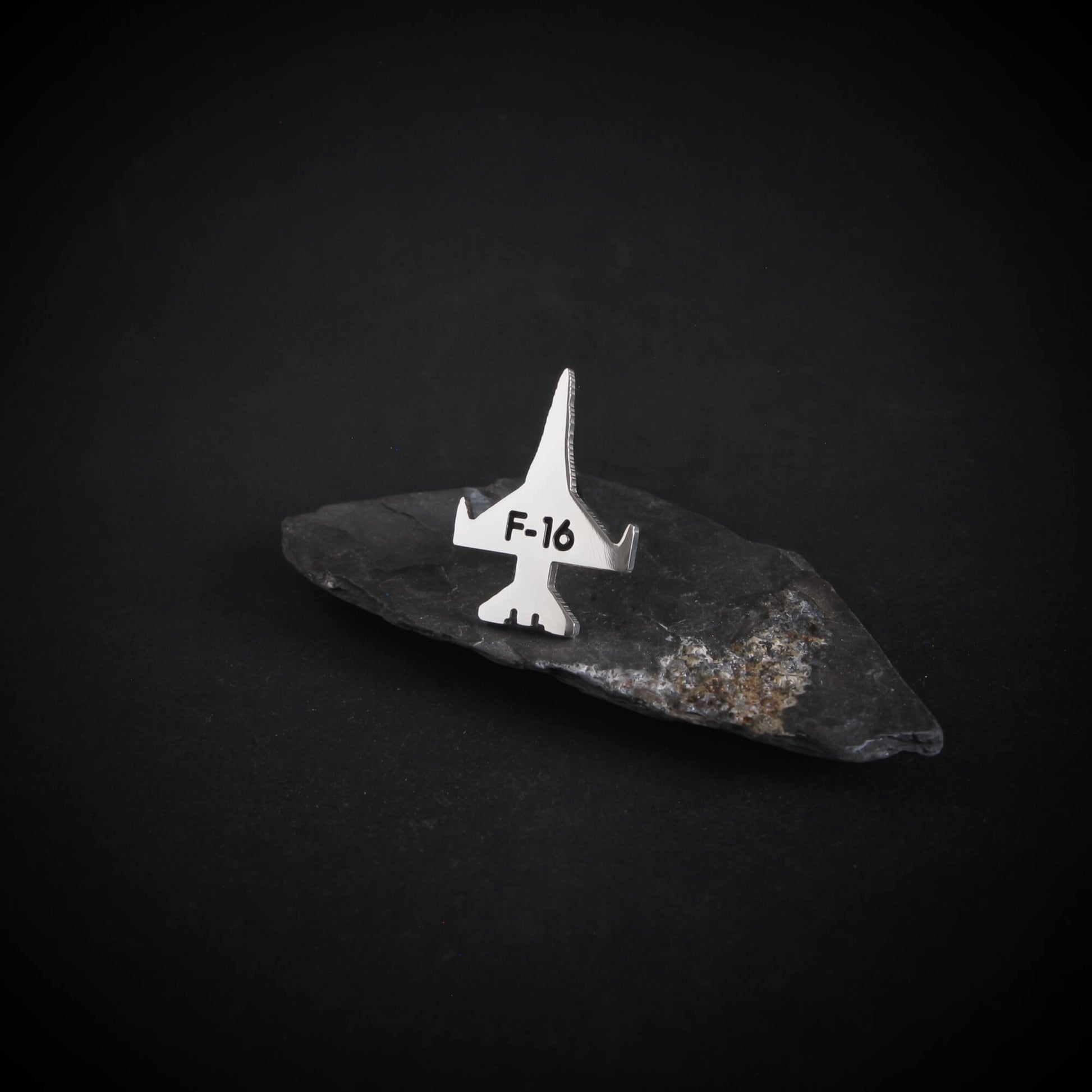 Silver F-16 Military jet fighter airplane tie tack on a dark stone surface with a black background