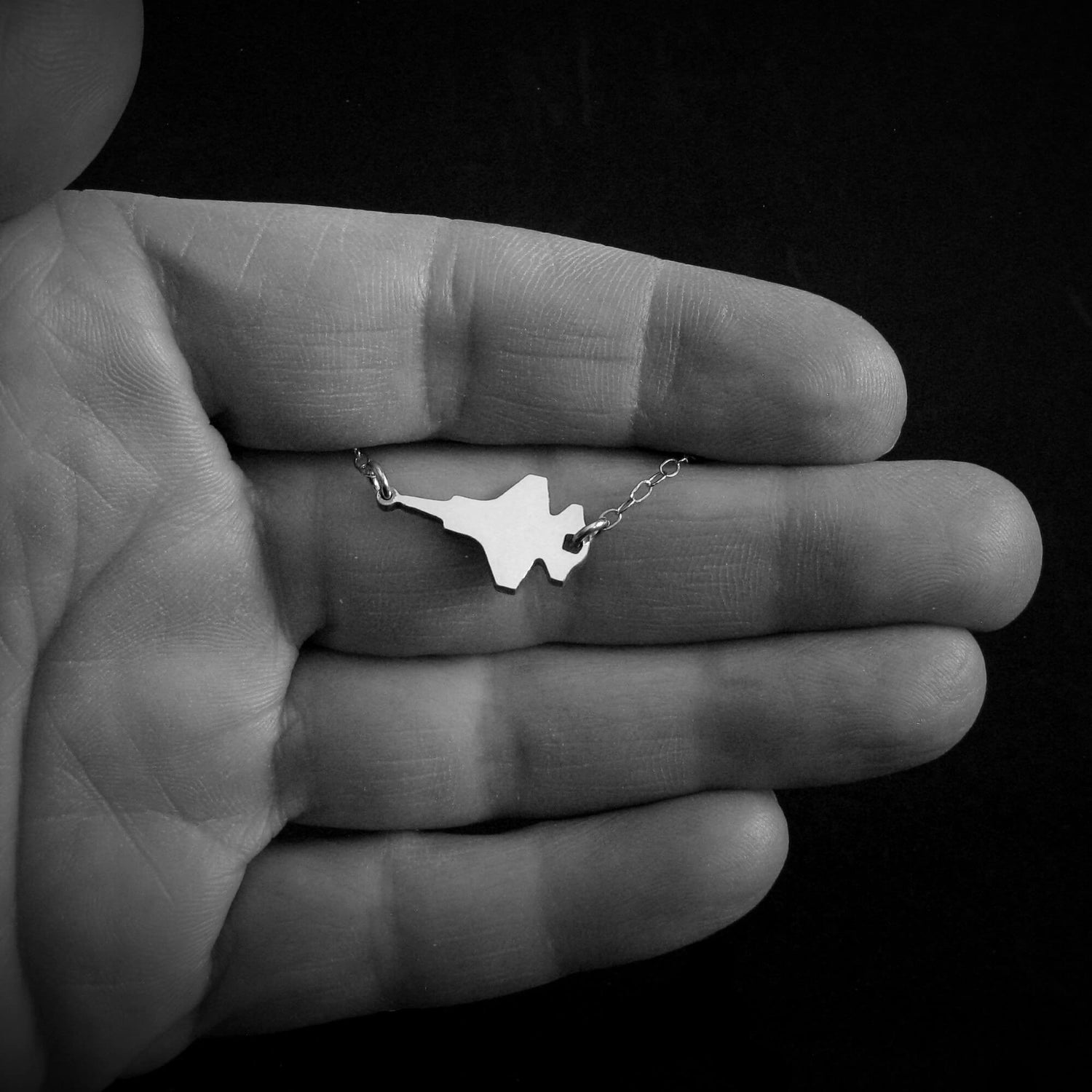 Small silver F-35 Military Fighter Jet necklace with a pendant held in a hand against a black background