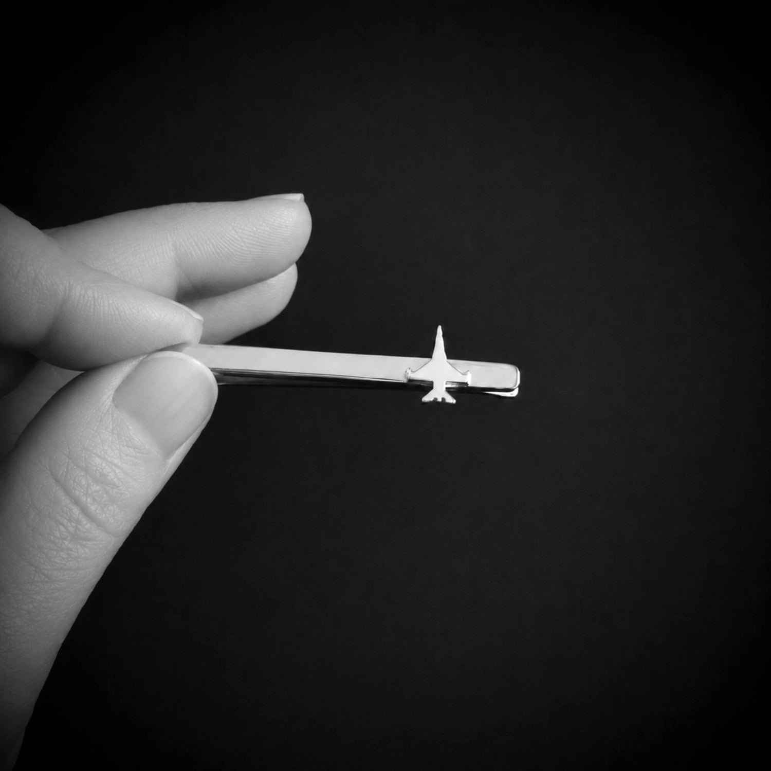 A hand holding a sterling silver tie bar with a small F 16, shown against a dark black background.