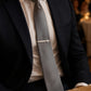 A close-up of a man wearing a navy suit jacket, white dress shirt, and a light gray tie with a sterling silver f 16 tie bar, set in a warm, blurred event environment.