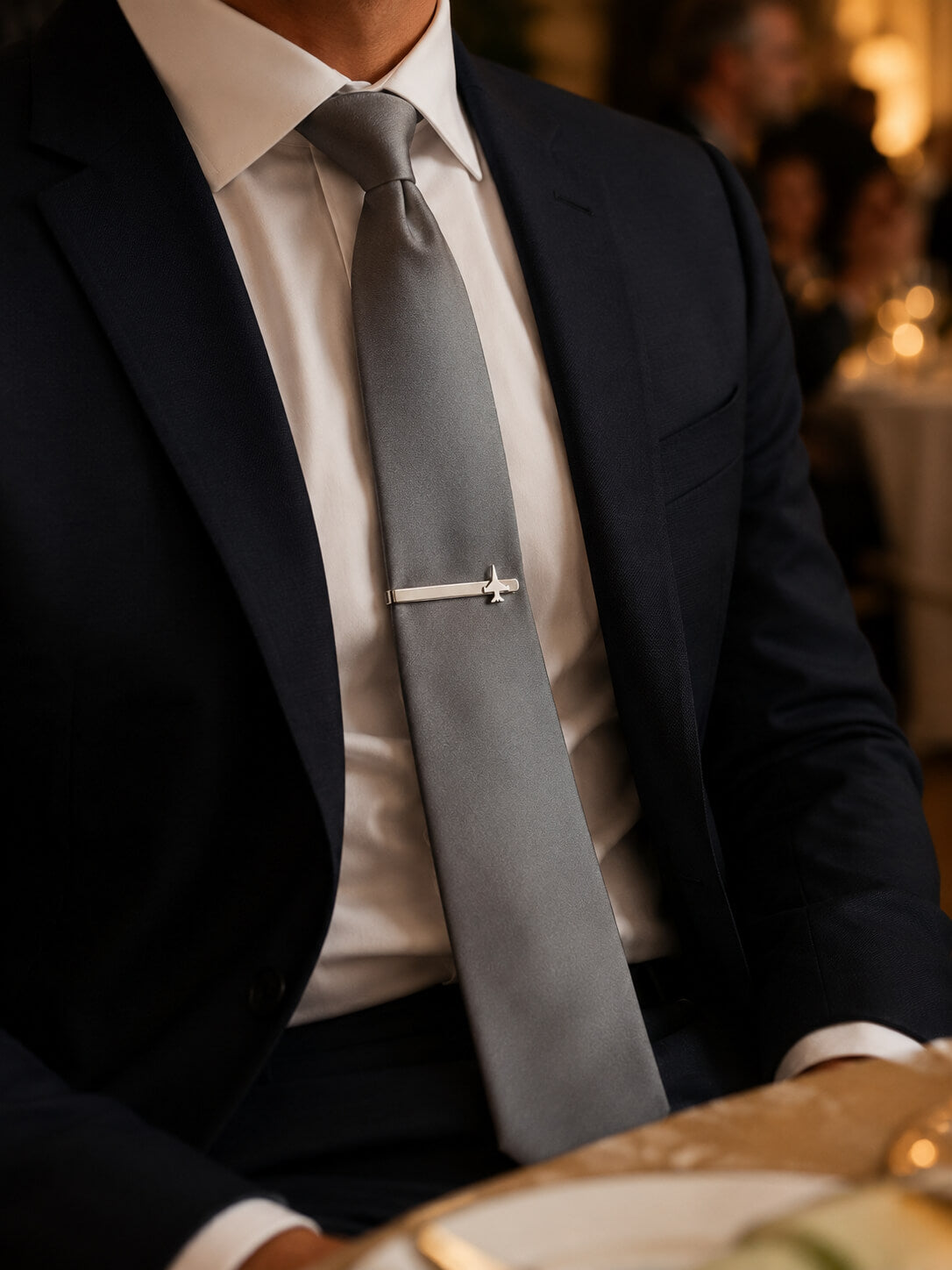 A close-up of a man wearing a navy suit jacket, white dress shirt, and a light gray tie with a sterling silver f 16 tie bar, set in a warm, blurred event environment.