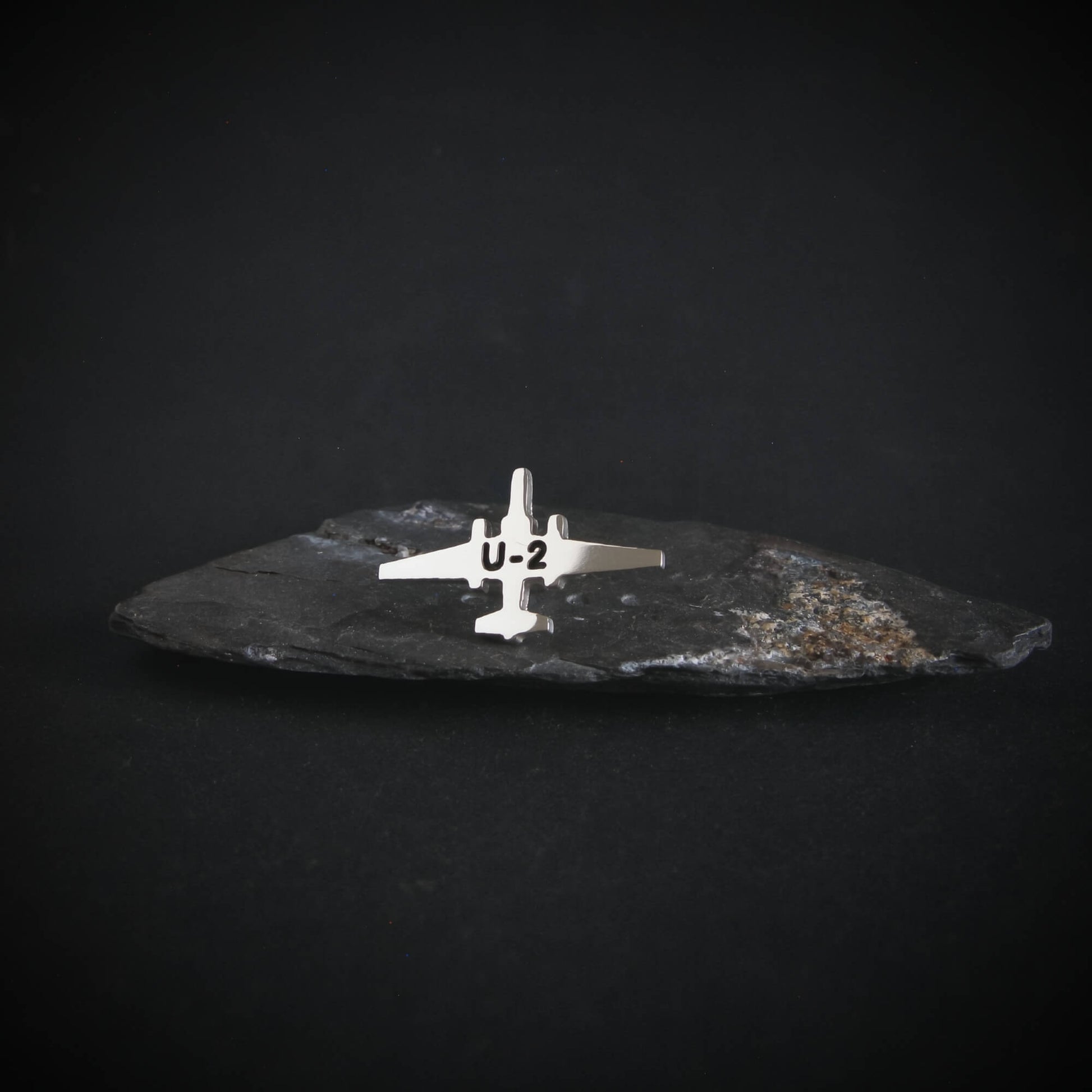 Silver U-2 spy plane tie tack on a dark stone surface with a black background