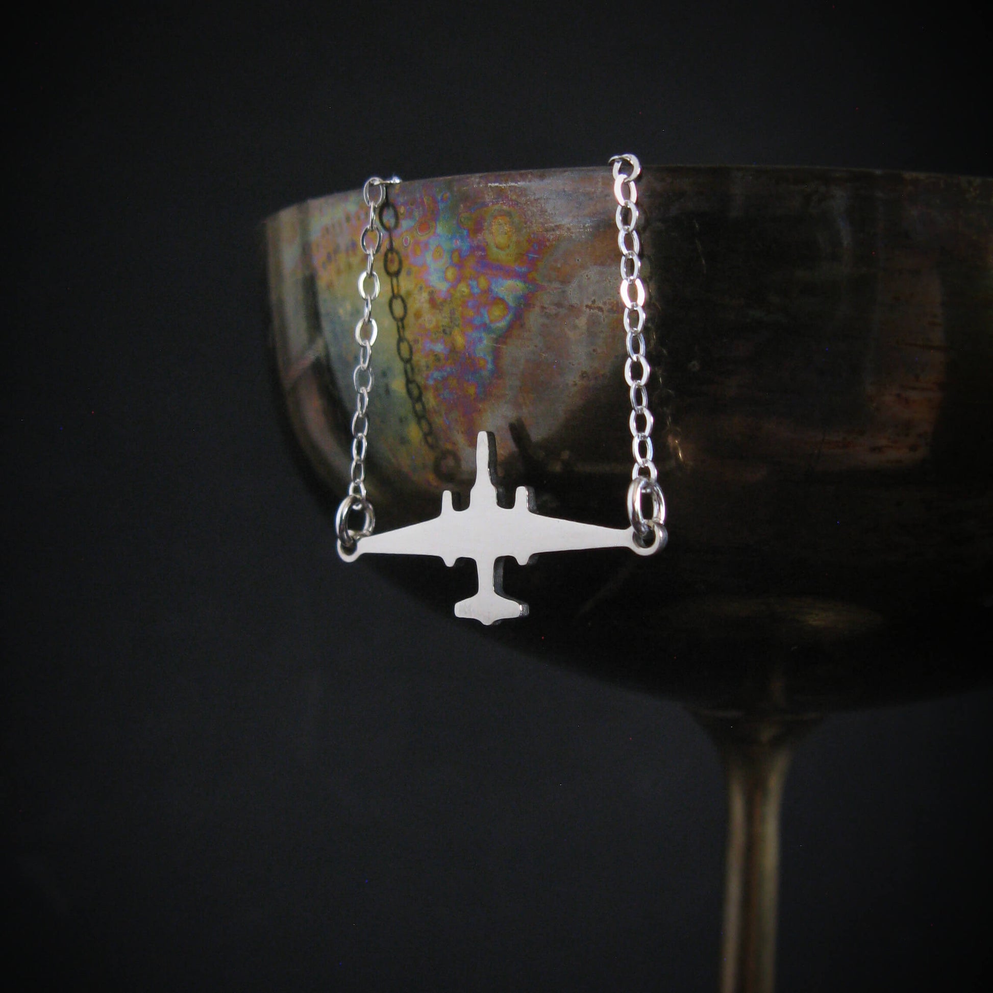 U-2 Dragon Lady military jet pendant necklace in sterling silver, draped over the rim of an iridescent metal wine glass against a dark background.